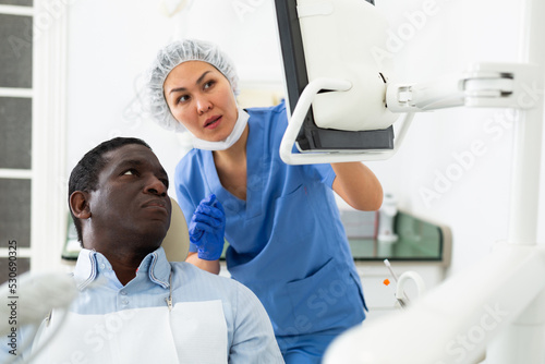 Qualified asian female dentist pointing at monitor explaining diagnosis to focused african american in dental office..