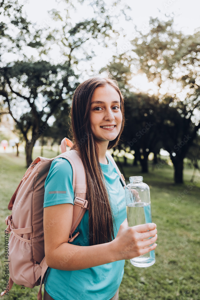 Teenage girl wearing turquoise t shirt and a pink backpack, holding a glass bottle with water in nature
