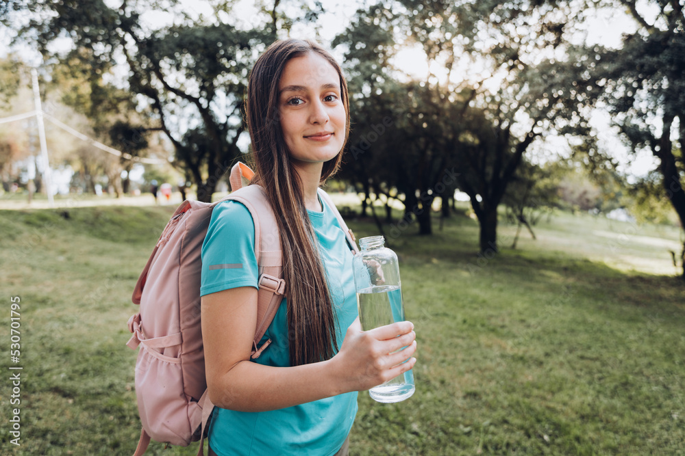 Teenage girl wearing turquoise t shirt and a pink backpack, holding a glass bottle with water in nature Teenage girl wearing turquoise t shirt and a pink backpack, holding a glass bottle with water in nature