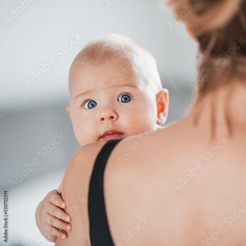 Portrait of sweet baby resting in mothers arms, looking at camera, touching mama shoulder. New mom holding little kid, embracing child with tenderness, love, care. Motherhood concept