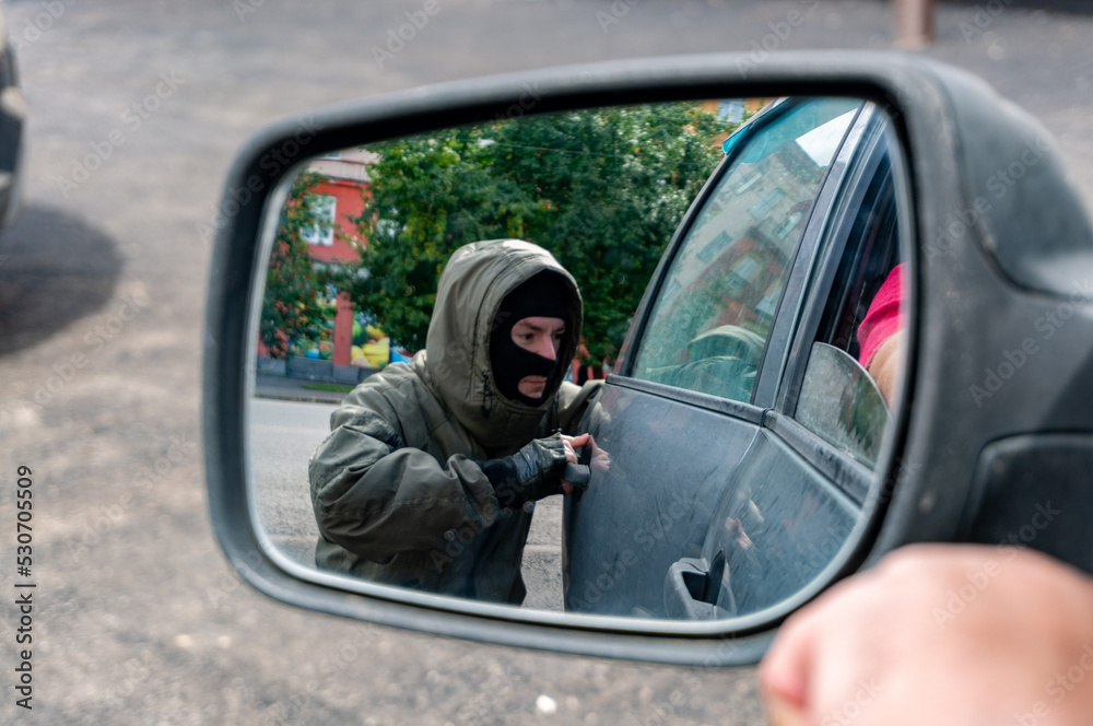 A masked street thief opens the back door of a car from a careless driver. View in the rear view mirror