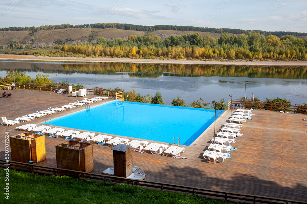 Obraz premium Blue pool without people on the riverbank in autumn. A great place to relax and unwind in nature. The pool and sun beds are waiting for visitors