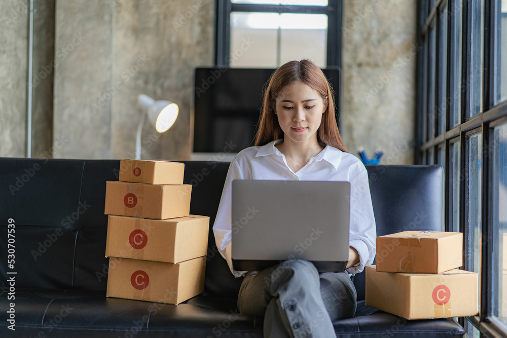 Asian woman sitting on sofa at home selling online with box and laptop receiving messages from customers sme business idea