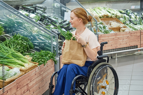 woman in a supermarket chooses vegetables, herbs, uses an eco-friendly bag, sitting in a wheelchair © evafesenuk