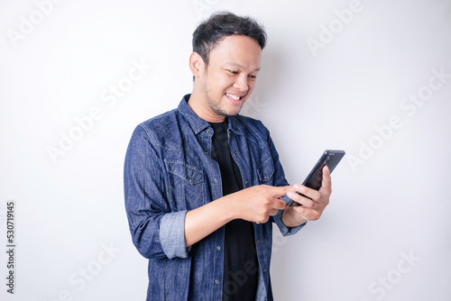 A portrait of a smiling Asian man is smiling and holding his smartphone wearing a navy blue shirt isolated by a white background
