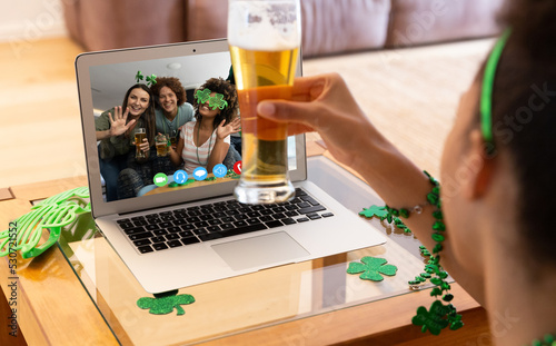 Mixed race woman holding beer having st patrick's day video call with friends on laptop at home