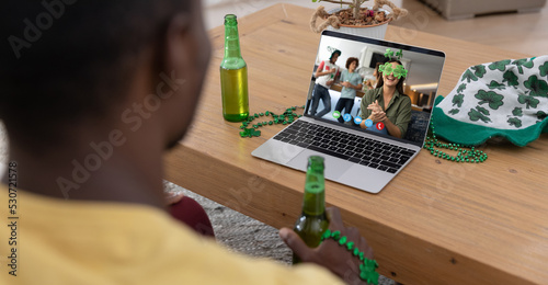 African american man holding beer making st patrick's day video call to friends on laptop at home