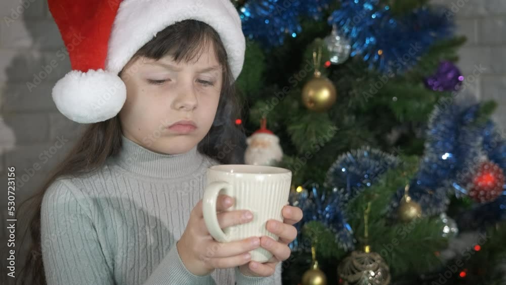 Little girl with hot milk at Christmas celebration. A stressed little girl in Santa hat celebrate the New Year with a mug of hot tea by the colorful fir-tree in the room. Little girl with hot milk at Christmas celebration. A stressed little girl in Santa hat celebrate the New Year with a mug of hot tea by the colorful fir-tree in the room.