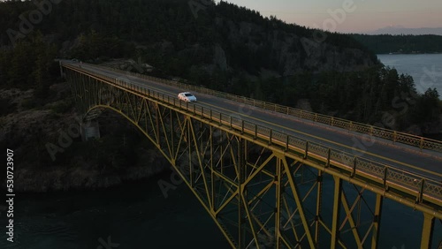 Static aerial shot of a white sedan driving across Deception Pass Bridge at sunrise.