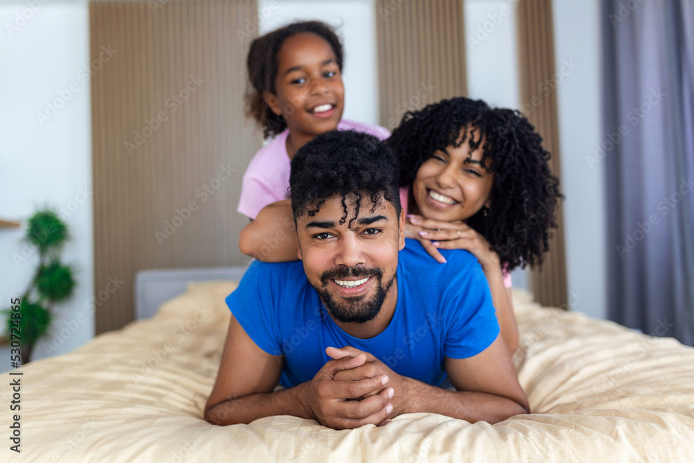 Portrait of beautiful young mother, father and their daughter looking at camera and smiling while lying on bed leaning on each other Portrait of beautiful young mother, father and their daughter looking at camera and smiling while lying on bed leaning on each other
