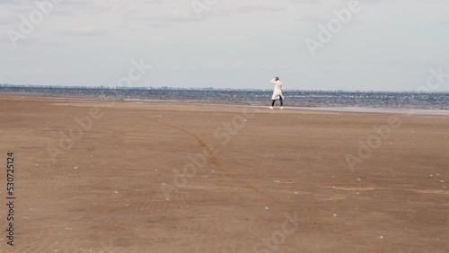 a beautiful woman with red hair in a white coat walks on a sandy beach on a cold sunny day, the unity of woman and nature