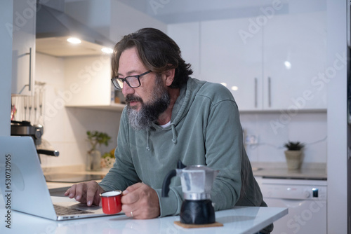 One man at home having breakfast before work drinking italian coffee. Modern people using computer laptop in the kitchen in house real lifestyle leisure. Male person indoor online activity at home