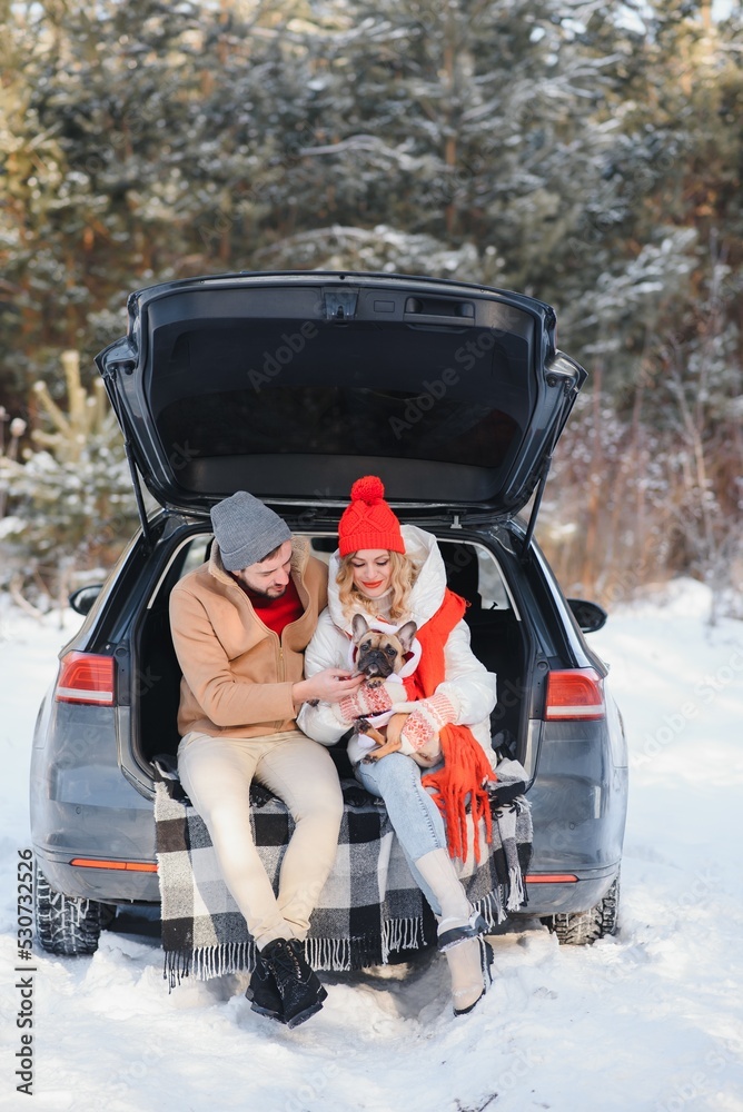 Smiling couple with dog sitting in open SUV car trunk in snowy forest. Enjoying each other in active winter holidays.