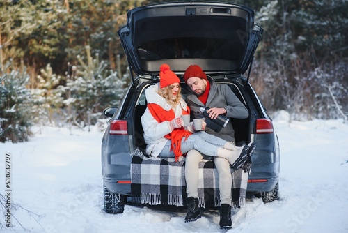 Smiling together. Nice couple have weekend at countryside at winter time. Sitting on the rear part of modern car.