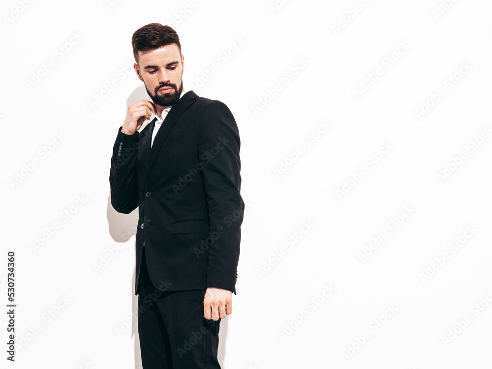 Portrait of handsome confident stylish hipster lambersexual model. Sexy modern man dressed in elegant black suit. Fashion male posing in studio near white wall. Isolated Portrait of handsome confident stylish hipster lambersexual model. Sexy modern man dressed in elegant black suit. Fashion male posing in studio near white wall. Isolated