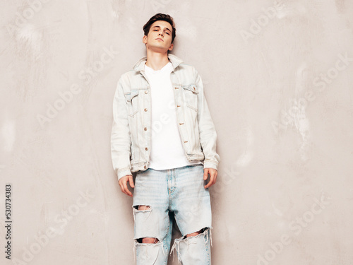 Portrait of handsome confident stylish hipster lambersexual model.Man dressed in jacket and jeans. Fashion male posing in studio near grey wall