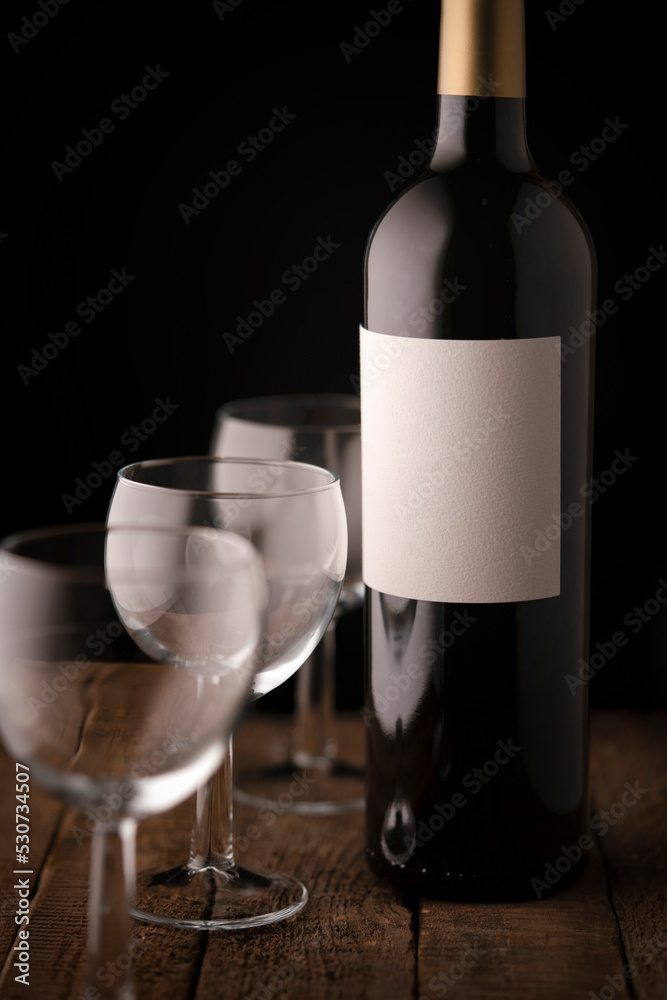 A bottle of red wine with an empty white label and three empty small wine glasses on a wooden table.