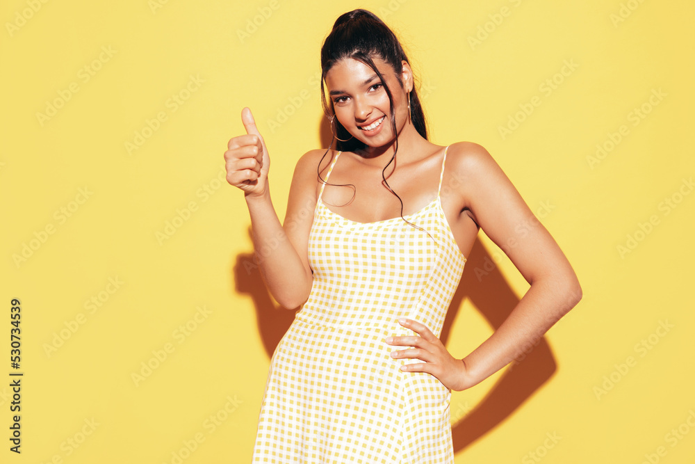 Young beautiful smiling female in trendy summer dress clothes. Sexy carefree woman posing near yellow wall in studio. Positive model having fun and going crazy. Cheerful and happy. Shows thump up Young beautiful smiling female in trendy summer dress clothes. Sexy carefree woman posing near yellow wall in studio. Positive model having fun and going crazy. Cheerful and happy. Shows thump up