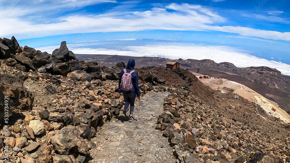 Rear view of hiking woman leaving the summit of volcano Pico del Teide on the island of Tenerife, Canary Islands, Spain, Europe. Island covered in cloud. Freedom concept. Scenic view volcanic terrain
