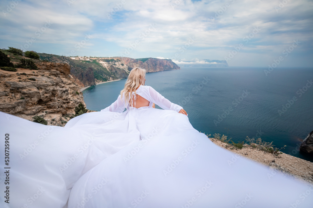 Blonde with long hair on a sunny seashore in a white flowing dress, rear view, silk fabric waving in the wind. Against the backdrop of the blue sky and mountains on the seashore.