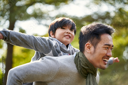 young asian father having fun carrying son on back outdoors in park