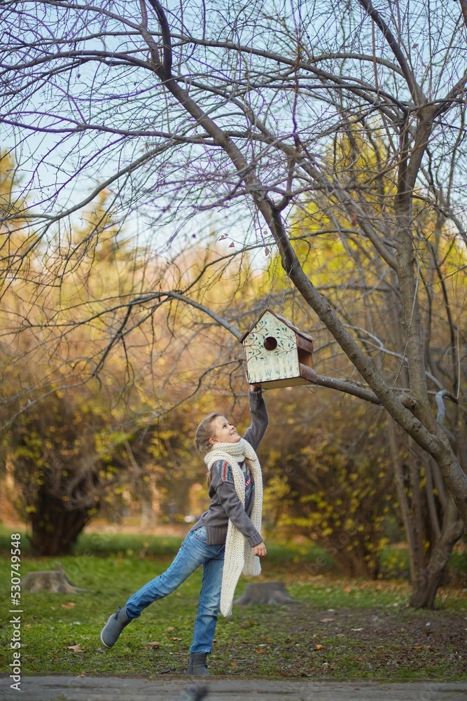 Obraz premium A girl in a knitted sweater in an autumn park looks at a birdhouse. September or October, yellow leaves on trees.