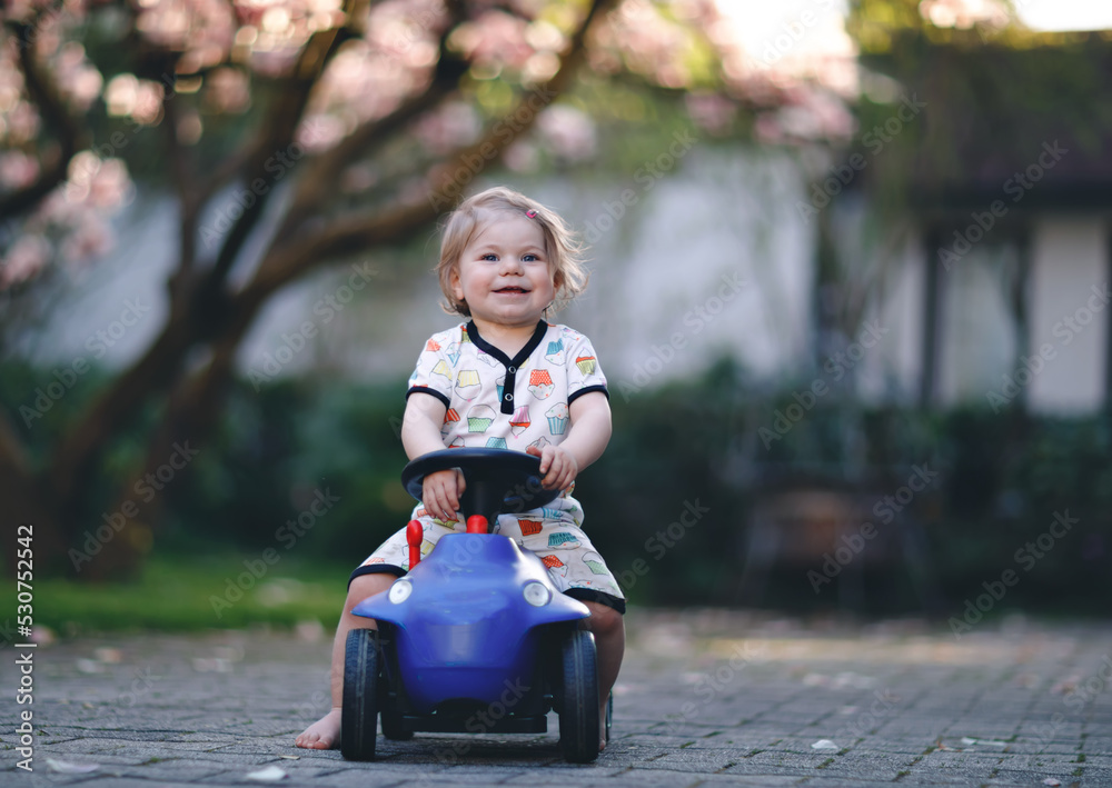 Cute little baby girl playing with blue small toy car in garden of home or nursery. Adorable beautiful toddler child with blossoming magnolia on background Cute little baby girl playing with blue small toy car in garden of home or nursery. Adorable beautiful toddler child with blossoming magnolia on background