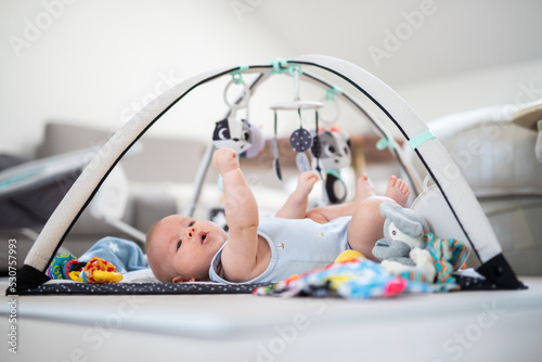 Cute baby boy playing with hanging toys arch on mat at home Baby activity and play center for early infant development. Baby playing at home