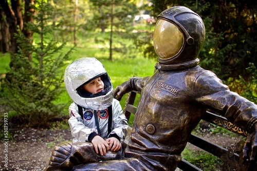 A young caucasian boy of five years old in a astronaut costume is sitting on the bench next to the statue of first astronaut Yuri Gagarin Translation from russian - Y.Gagarin