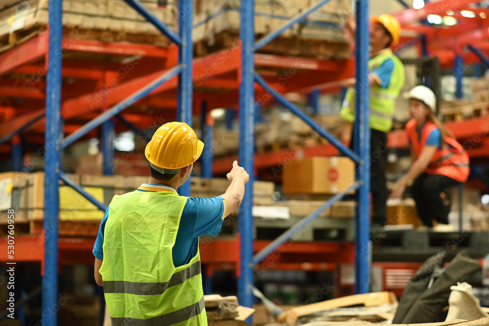 Back view of male manager wearing hardhat and reflective jacket pointing at upper shelf with goods in warehouse Back view of male manager wearing hardhat and reflective jacket pointing at upper shelf with goods in warehouse