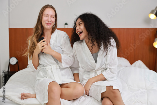 Young happy diversity ethnic beautiful woman in bathrobe sit on bed and playing social media on mobile smart phone and having fun together. Women lesbian couple daily LGBTQ lifestyle and love concept.