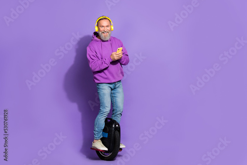 Photo of senior healthy cheerful man dressed purple hoodie on monowheel typing message smartphone isolated on violet color background photo