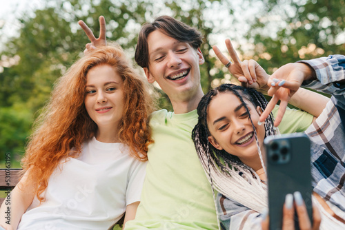 Front view of three friends sitting on the wooden bench in a park taking a selfie using a modern device. Selfie of young smiling people having fun together