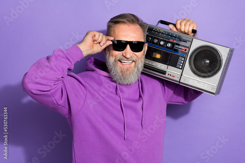 Photo of old handsome good mood man wear purple hoodie fingers touch sunglass hold boombox on shoulder isolated on violet color background