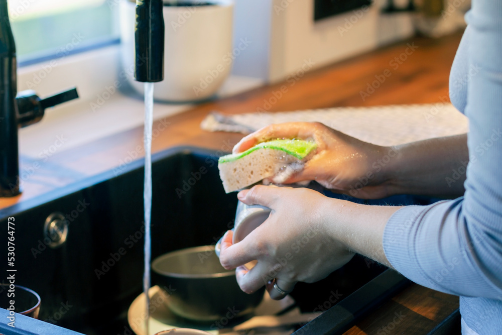 A woman is washing dishes in the kitchen. Selective focus on the hand. Cup and washing sponge in the hand. Kitchen with window and street view.