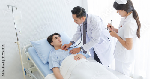 Senior doctor and young male patient who lie on the bed while checking symptom, consult and explain with nurse taking note and supporting in hospital wards.