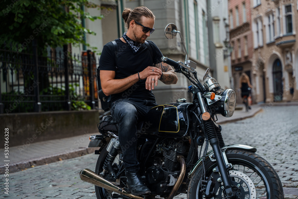 Shot of cool man biker with tattooes hand looking at watches riding bike at streets.