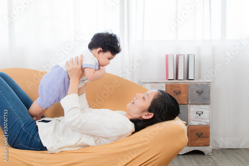 Selective focus healthy asian mother play and tease with newborn baby in living room, mom holding lifting toddler on brown sofa with fun and tender. Adorable little boy having fun with mom togerther. photo