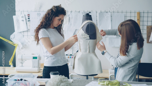 Creative fashion designers are pinning cut out pieces of fabric to mannequin while sewing women's garment in modern light studio. Ladies are concentrated on their work.