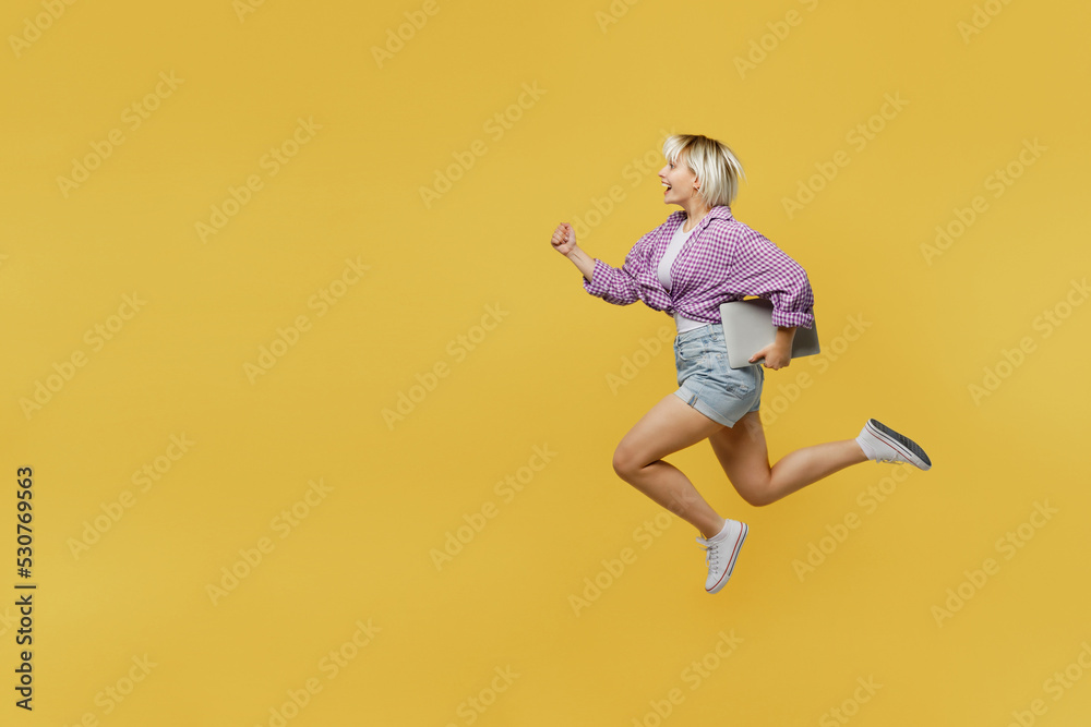 Full bodyside view young blonde IT woman 20s she wear pink tied shirt white t-shirt jump high run fast hurry up hold use work on laptop pc computer isolated on plain yellow background studio portrait.