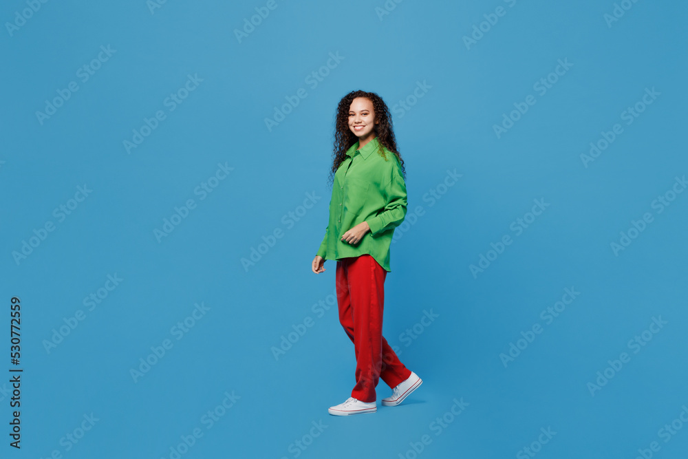 Full body side view cheerful young woman of African American ethnicity 20s she wear green shirt walking going look camera isolated on plain blue background studio portrait. People lifestyle concept. Full body side view cheerful young woman of African American ethnicity 20s she wear green shirt walking going look camera isolated on plain blue background studio portrait. People lifestyle concept.