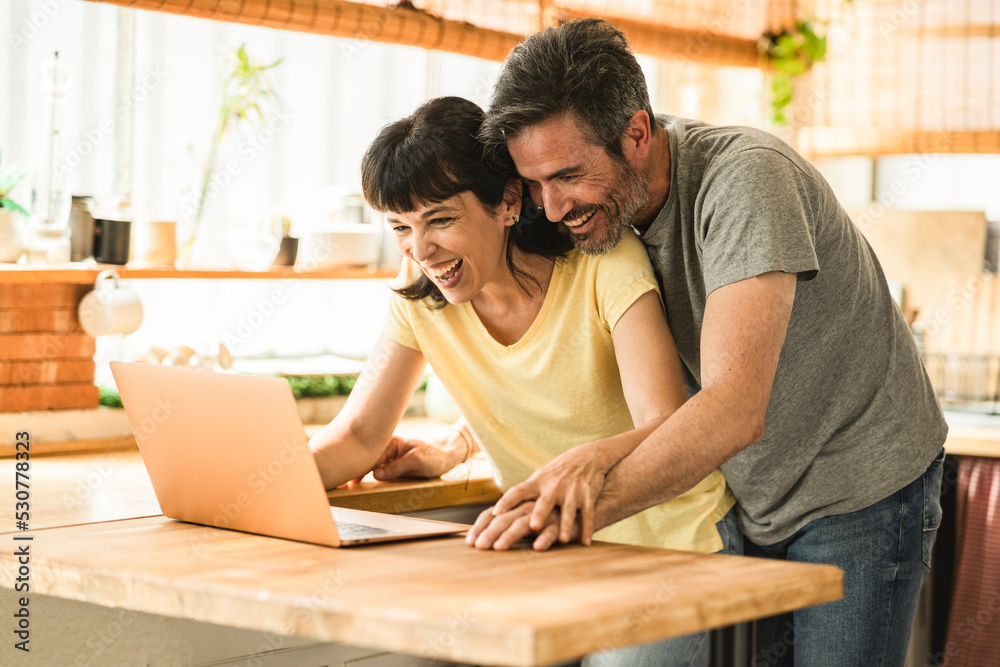 Smiling mature couple in the kitchen at home and having fun shopping online on the computer together. Smart modern happy man and woman looking at laptop screen paying with credit card or web. Smiling mature couple in the kitchen at home and having fun shopping online on the computer together. Smart modern happy man and woman looking at laptop screen paying with credit card or web.
