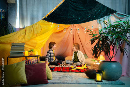 Two young cute boys are playing inside a nice comfy tent that has a hippie vibe to it, the tent is an indoor one photo