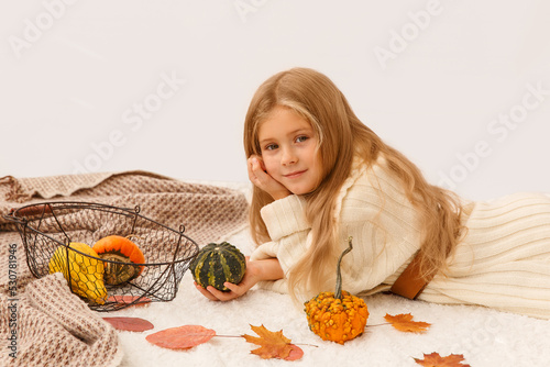 Blonde girl in white sweater holding pumpkin with thanksgiving symbol. Blanket and little pumpkin on the floor. Kidis laying on the floor photo