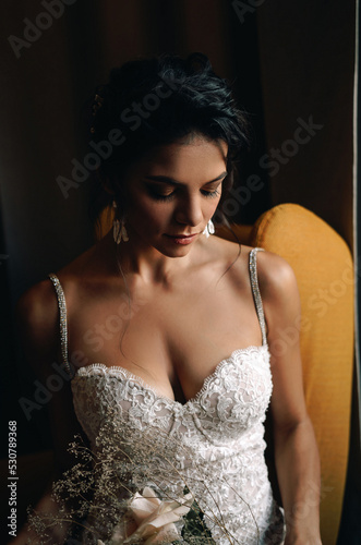 Brunette girl bride on a yellow armchair with a bouquet. Stylish wedding look at the hotel.