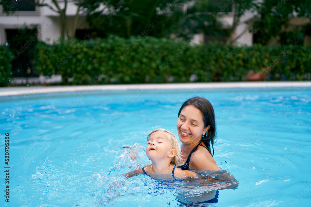 Little girl swims in the pool with her head raised above the water, supported by her mother. High quality photo