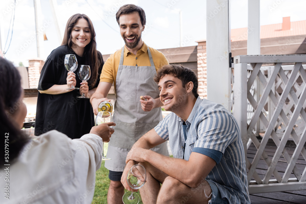 Positive man pouring wine near interracial friends in backyard