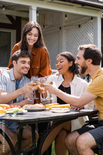 Cheerful multiethnic friends toasting with wine near grilled food during picnic outdoors