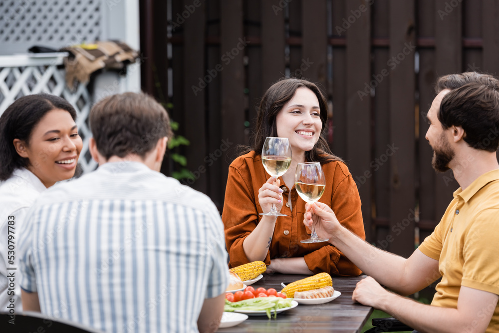 Positive people holding wine while blurred interracial friends talking near tasty food outdoors Positive people holding wine while blurred interracial friends talking near tasty food outdoors