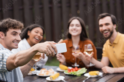 cheerful man taking selfie with blurred multiethnic friends during bbq party